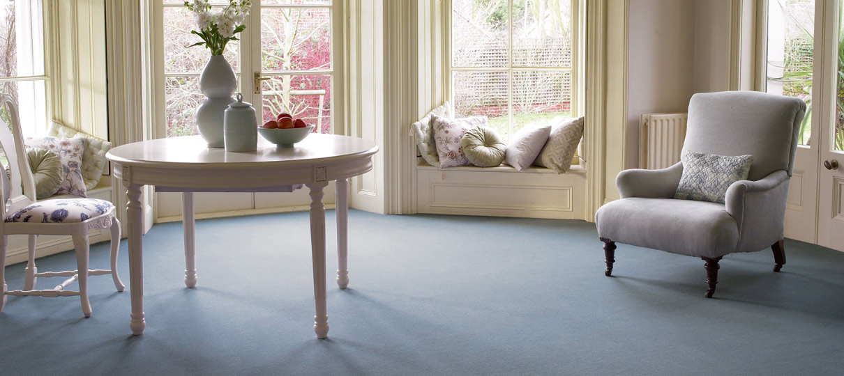 Pale blue carpet running through a bay-window sitting room, with a white painted table and armchair.
