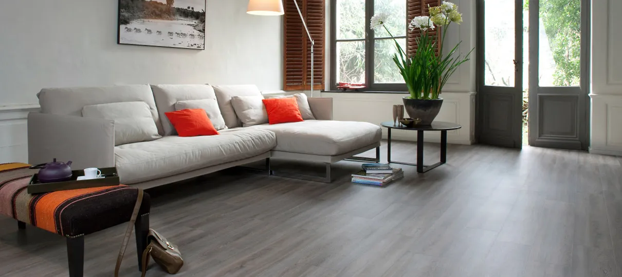 Grey wood-effect plank flooring in a bright living room with a cream corner sofa and tall shuttered windows.