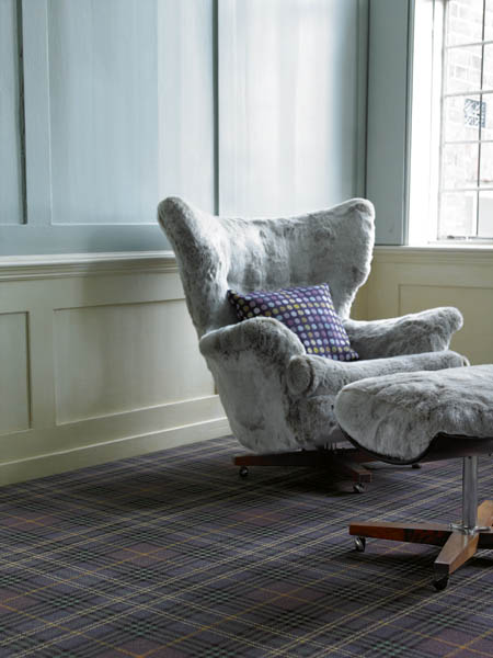 Purple and grey tartan carpet in a panelled room, with a grey sheepskin armchair and footstool.
